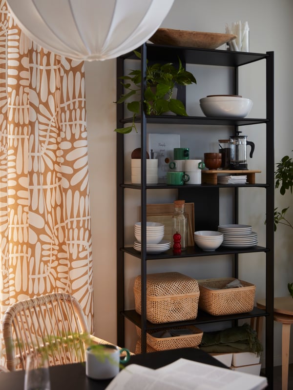 A black JÄTTESTA shelving unit with tableware and SMARRA boxes by a wall near a dining area with a black LISABO table.