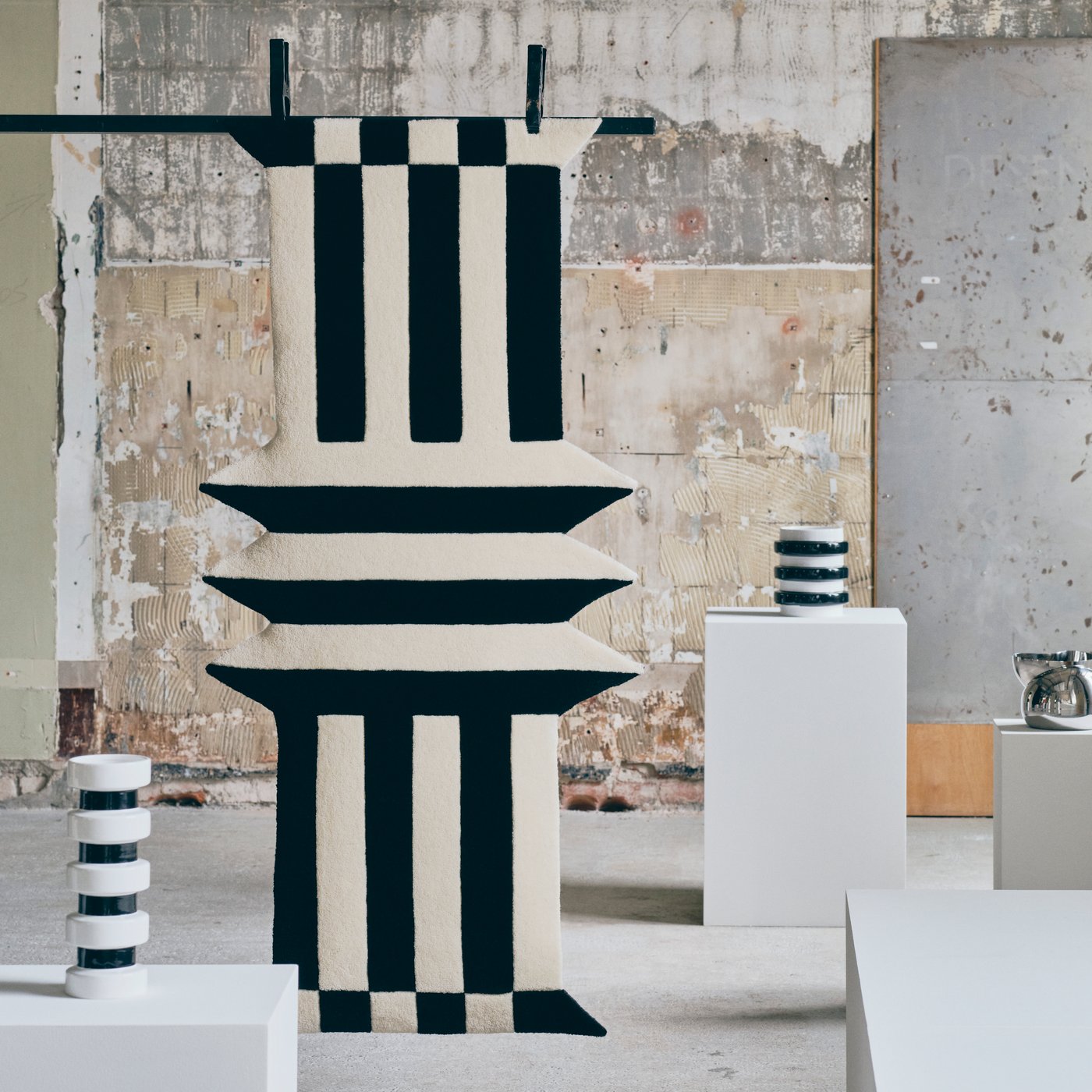 A gallery style room where three black and white objects are showcased, in the middle a large black and white rug.