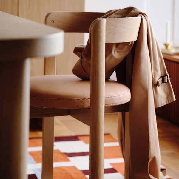 A STOCKHOLM chair in oak and leather with a garment hanging over he backrest, beside a STOCKHOLM tale in oak veneer and rug.