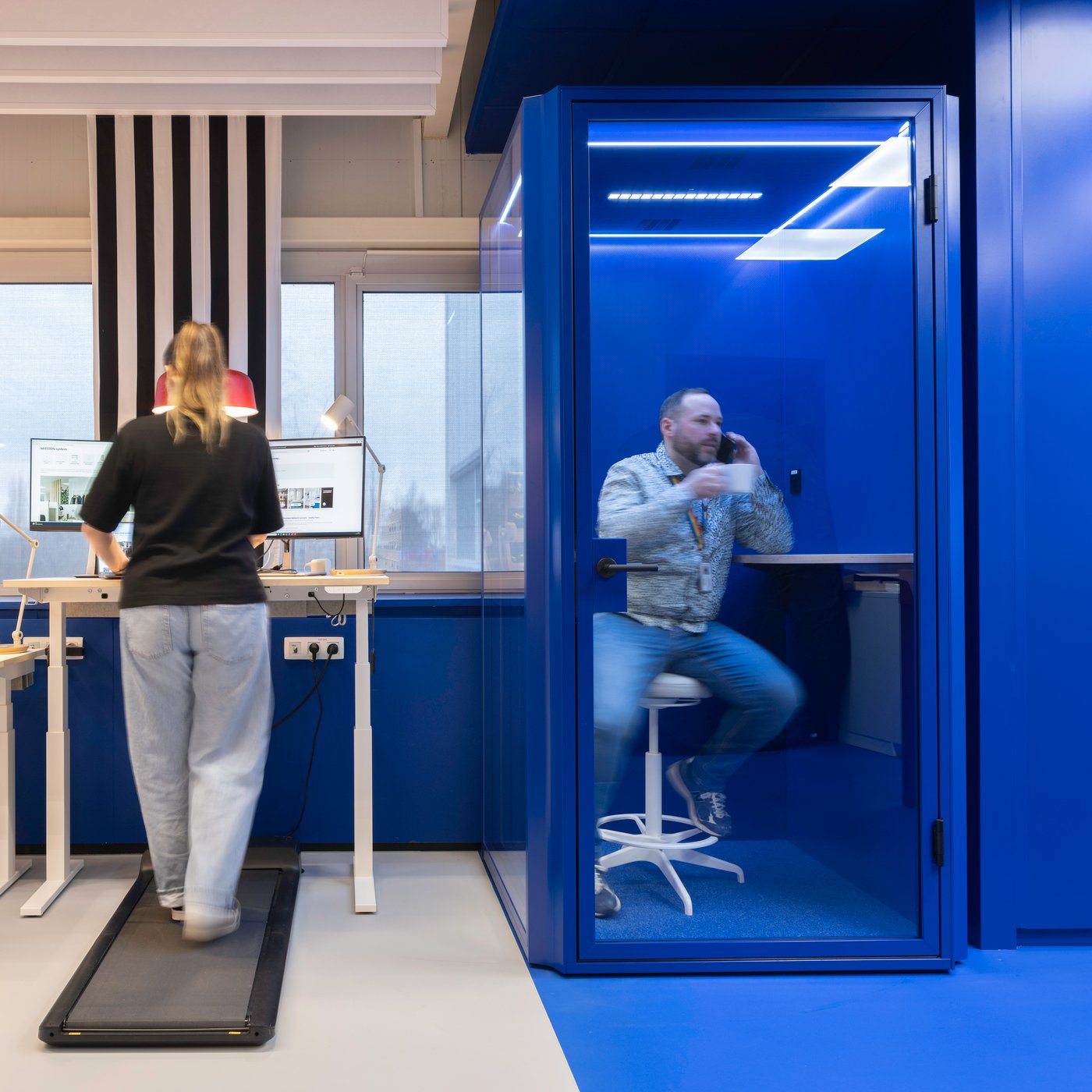 Au premier plan, une personne marche sur un bureau-tapis de marche tout en regardant un écran d’ordinateur. À sa droite, une autre personne est assise dans une cabine acoustique transparente bleu vif, en pleine conversation téléphonique. À l’arrière-plan, on distingue des bureaux avec ordinateurs et équipement de bureau, dans un ensemble dominé par des tons de bleu et de blanc, avec quelques touches de noir et de rouge.