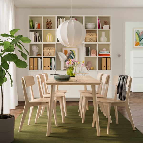 A cozy dining area features a wooden table, six chairs, a vase of flowers, and a lamp, with shelves filled with decor and books in the background.