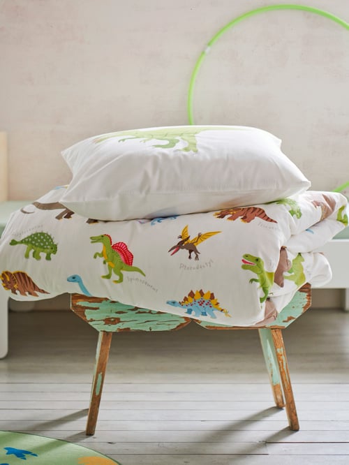 Ein Hocker mit gefalteter Bettdecke und Kissen mit JÄTTELIK Bettwäsche mit einem bunten Dinosauriermuster.