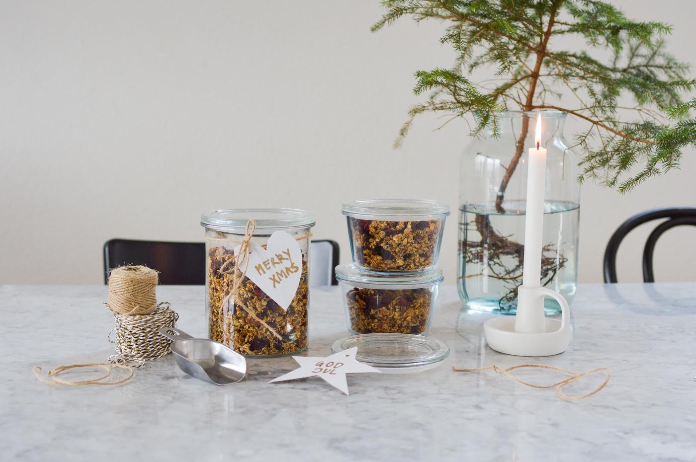 Festliche Szene mit Gläsern voller Müsli mit der Aufschrift „Merry Xmas“, Schnur und einer Metallschaufel auf einem Marmortisch. Eine brennende Kerze und ein kleiner Baum sorgen für Gemütlichkeit.