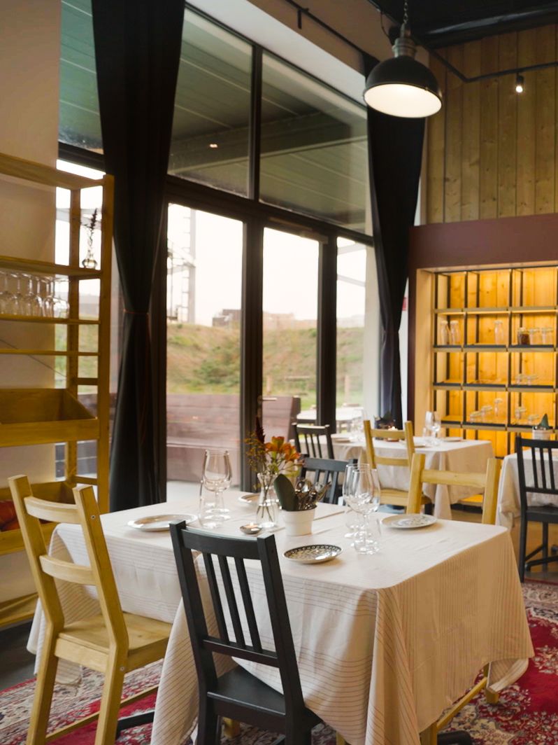 Gezellig restaurant met witte tafels, bloemen, glazen en houten stoelen. Grote ramen met zicht naar buiten en warme houten accenten binnen.