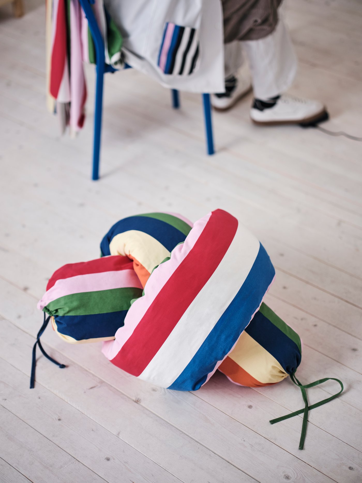 A tie-knot pillow lies on a bright wooden floor. It’s made in the candy-colored striped pre-cut fabric called JÄTTEVÄDD.