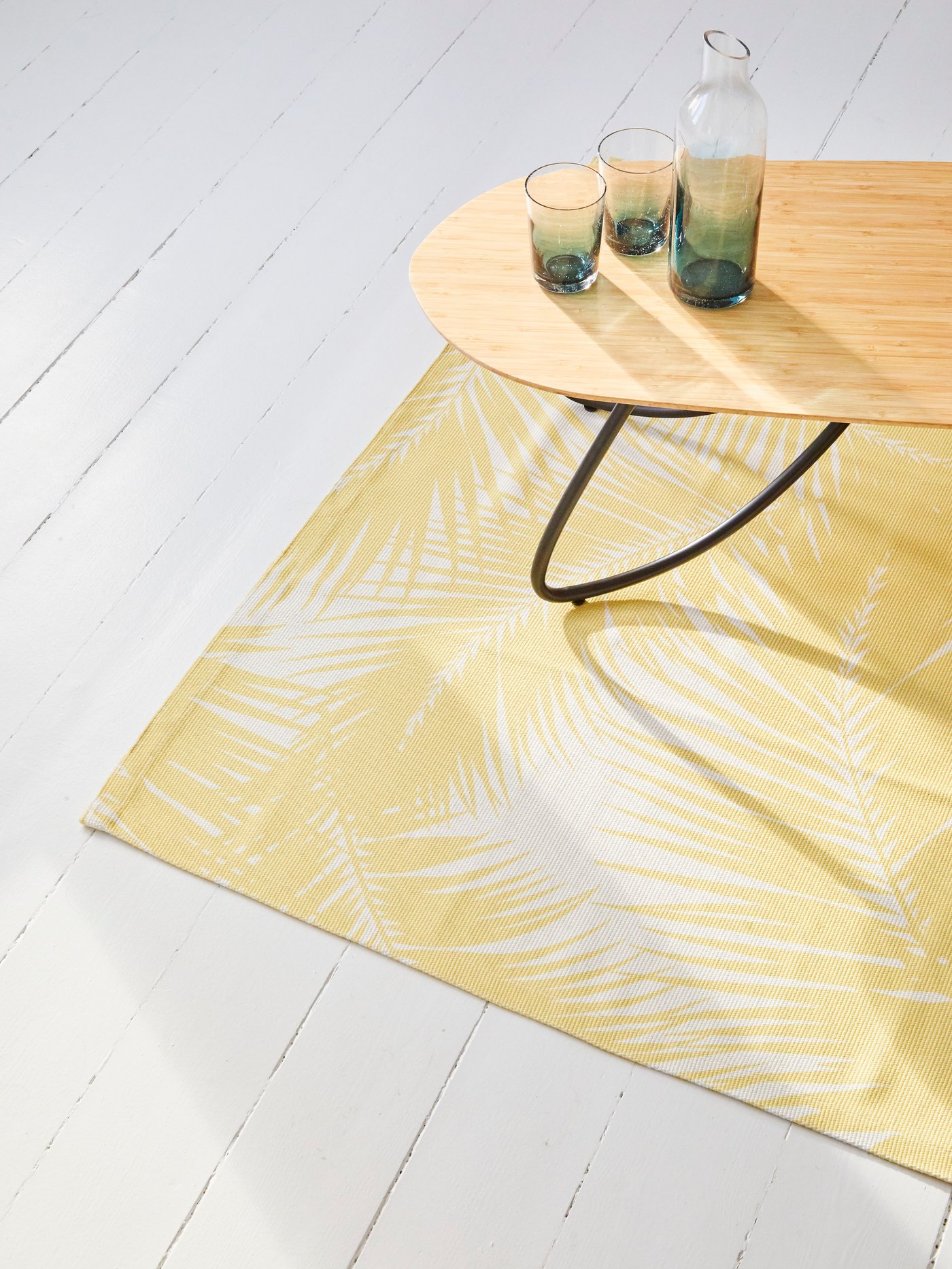 A coffee table in bamboo holding a carafe and glasses, standing on KÅSEBERGA rug with a yellow and white palm-inspired print.
