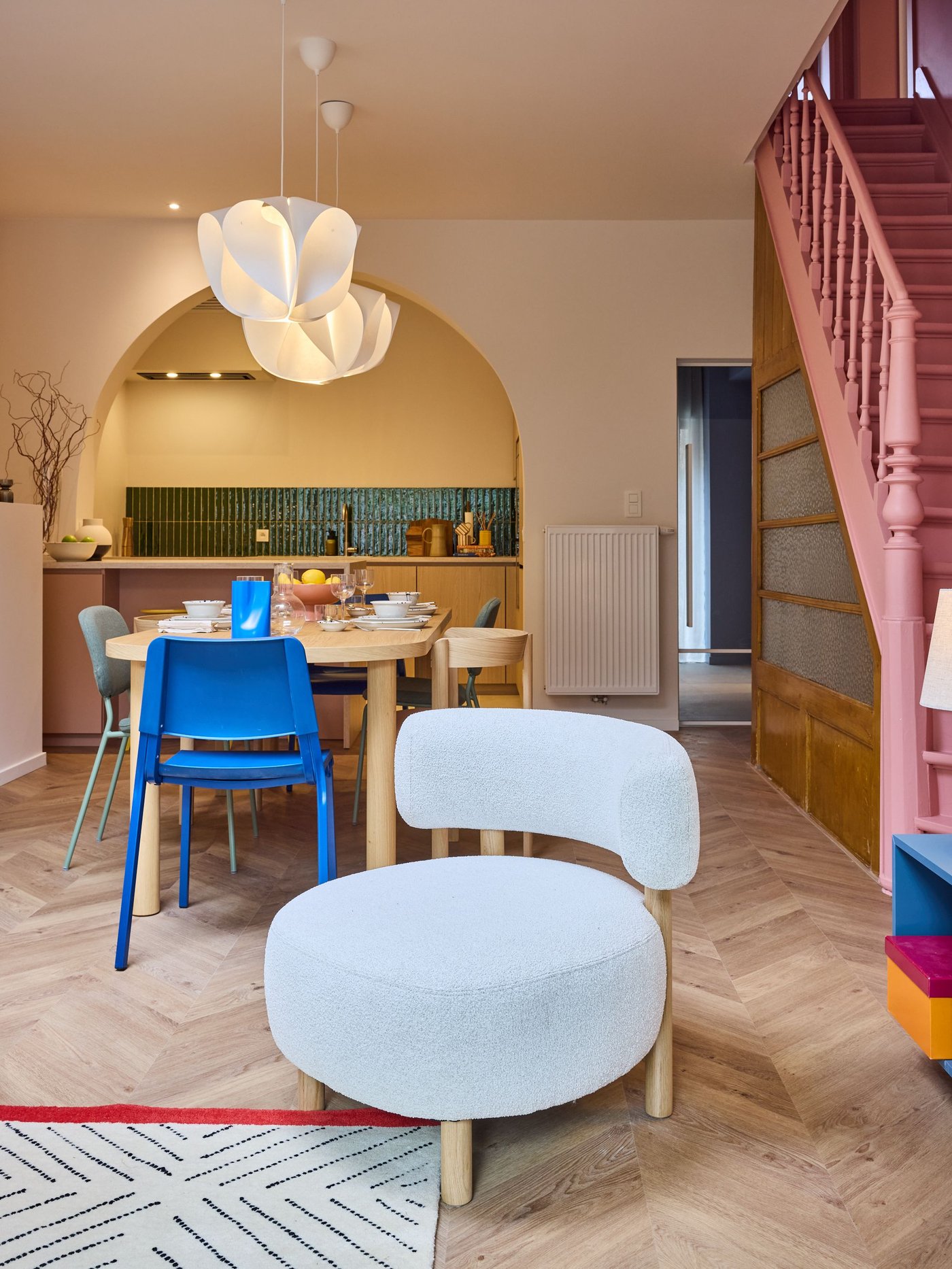 Eetruimte met houten tafel en blauwe stoelen, boog naar keuken, witte hanglamp en roze trap, lichte houten vloer en warm interieur.