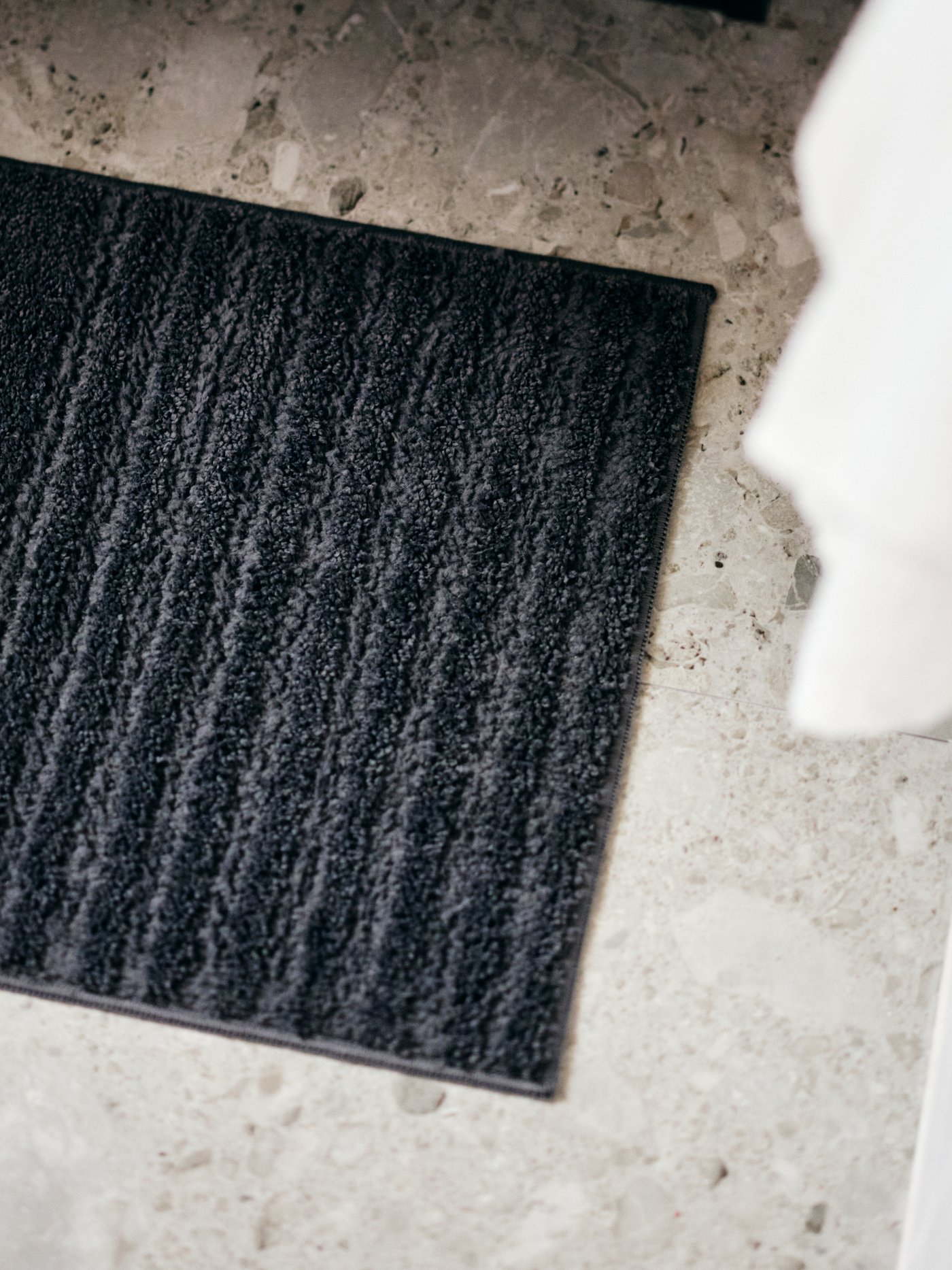 A bathroom corner with hanging towels and a dark-grey VINNFAR bathroom mat on the tiled floor.