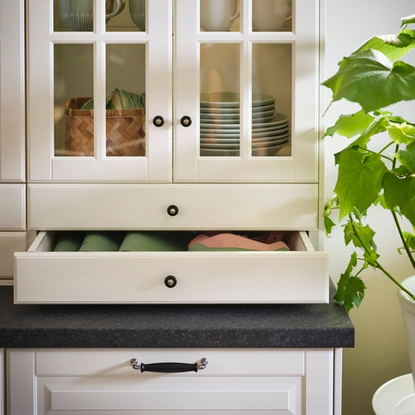 A cabinet with off-white BODBYN glass doors and drawer fronts displays plates and cups, and neatly folded linens.