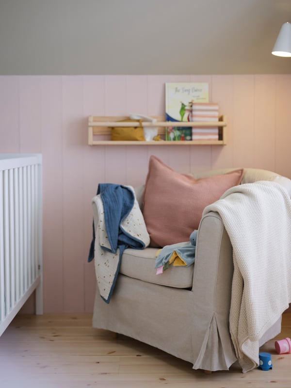 A TULLSTA armchair with a Lofallet beige cover, blankets hanging on the arms and a cushion in a pink KLOTSTARR cover on it.