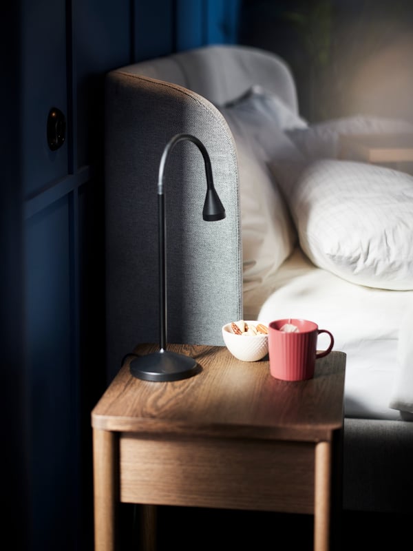 La tête de lit d’un lit matelassé TUFJORD flanqué d’une table de chevet TONSTAD noire sur laquelle se trouve une lampe de bureau à DEL NÄVLINGE noire.