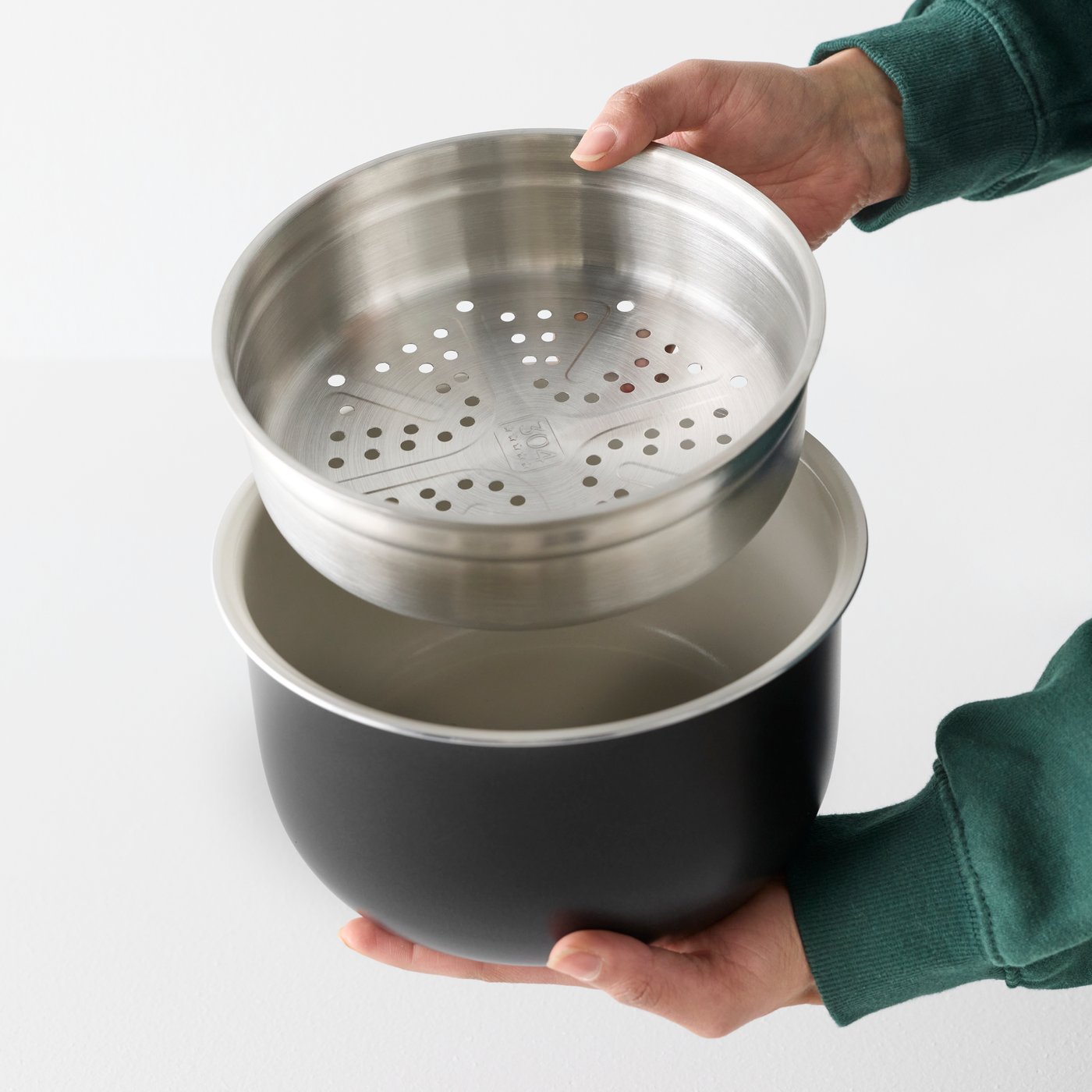 A close-up of hands holding a black TILLREDA multicooker featuring a stainless steel demonstrating the steaming insert on top.