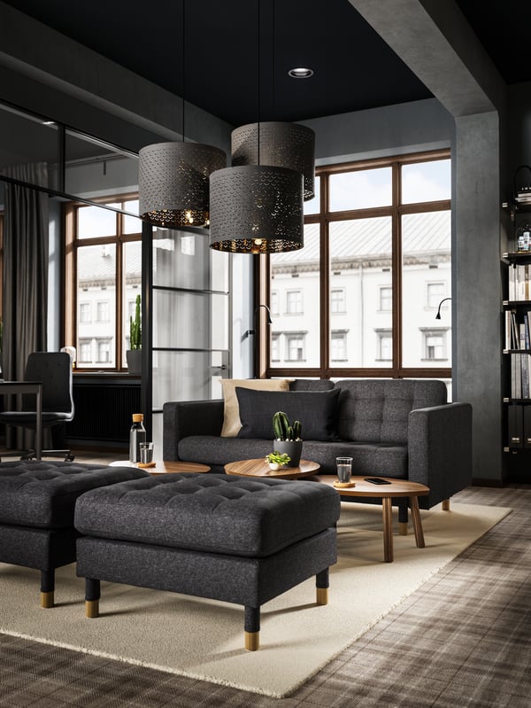 A lounge area created with a dark LANDSKRONA sofa and footstools with small wooden tables and three hanging lamps.