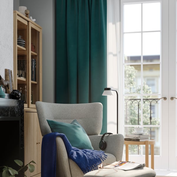 Ein beige grauer OSKARSHAMN Ohrensessel im Wohnzimmer. Am Fenster eine Kombination aus hellen HILJA Gardinen mit grünen Sanela Vorhängen.