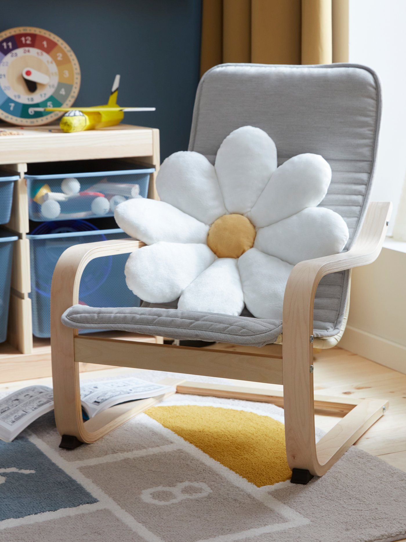 Prästkrageformad kudde ligger i en beige POÄNG barnfåtölj vid ett fönster i ett barnrum med förvaringslösningar.
