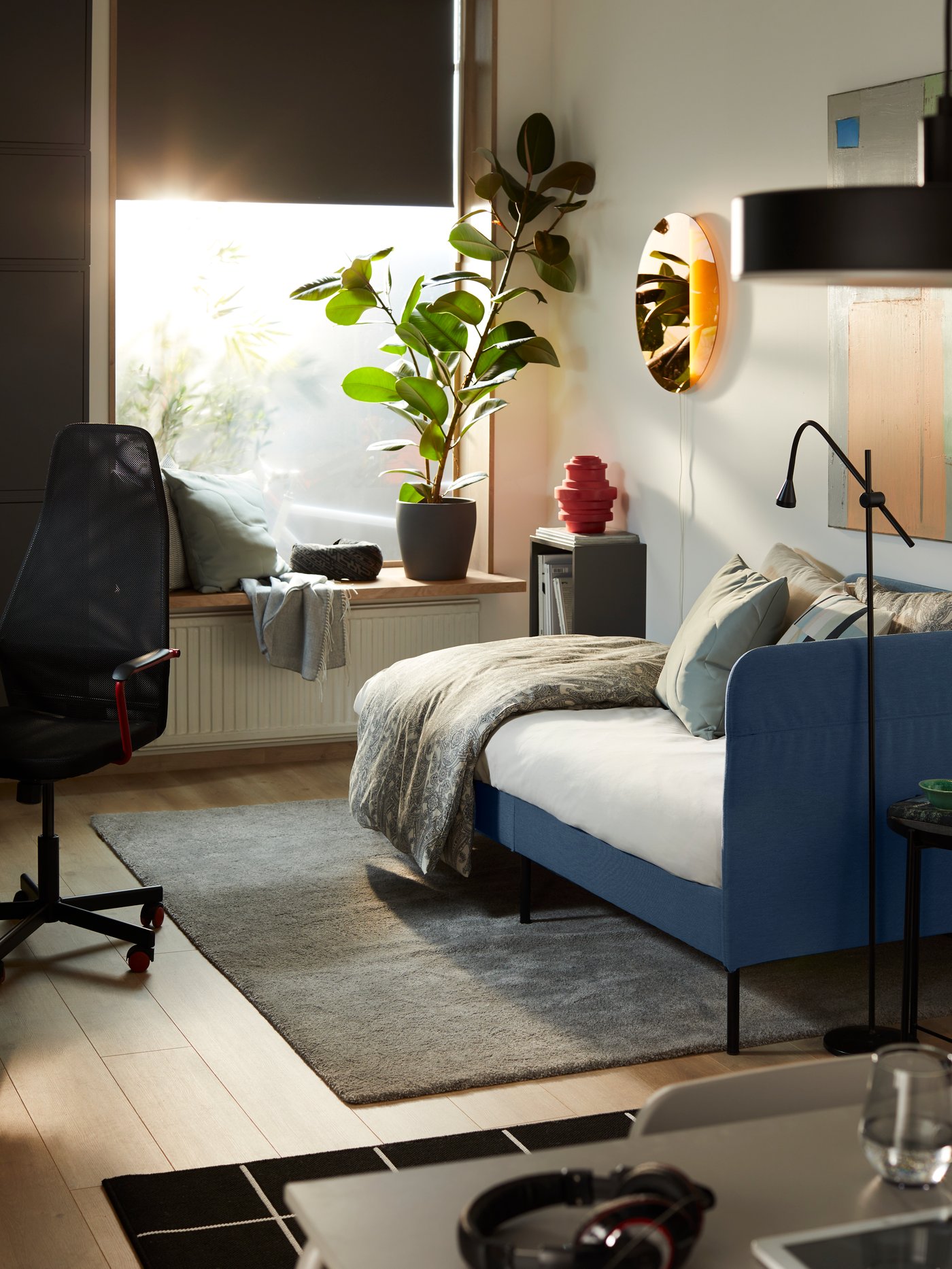 Een laagstaande zon schijnt in een opgeruimde kamer met een BLÅKULLEN bed, een bureau, een zwarte HUVUDSPELARE gamestoel, een plant en decoraties.