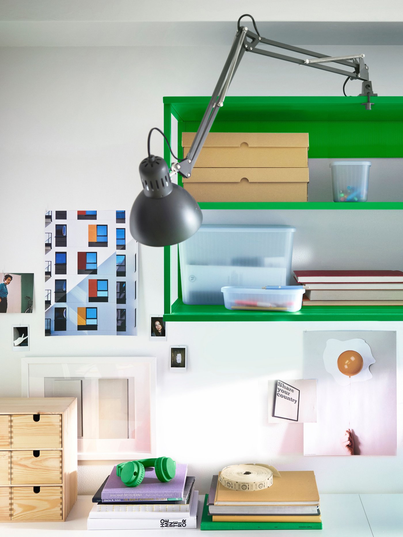 Workspace with a green wall shelf holding boxes and containers, a gray adjustable desk lamp, books, and colorful artwork on the wall.
