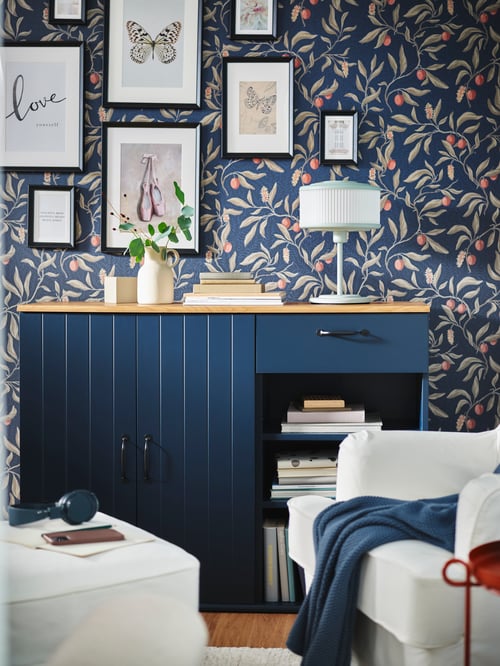 A blue SKRUVBY sideboard is against a wall with floral-themed wallpaper and framed pictures. A white armchair is in front.