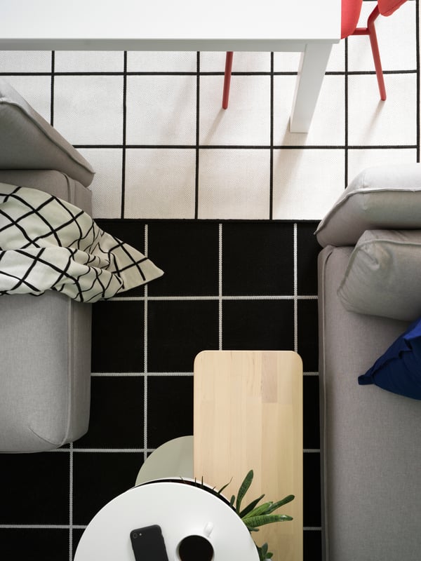 A view from above where you see two rugs in a black and white square pattern, a white dining table, and a grey sofa.