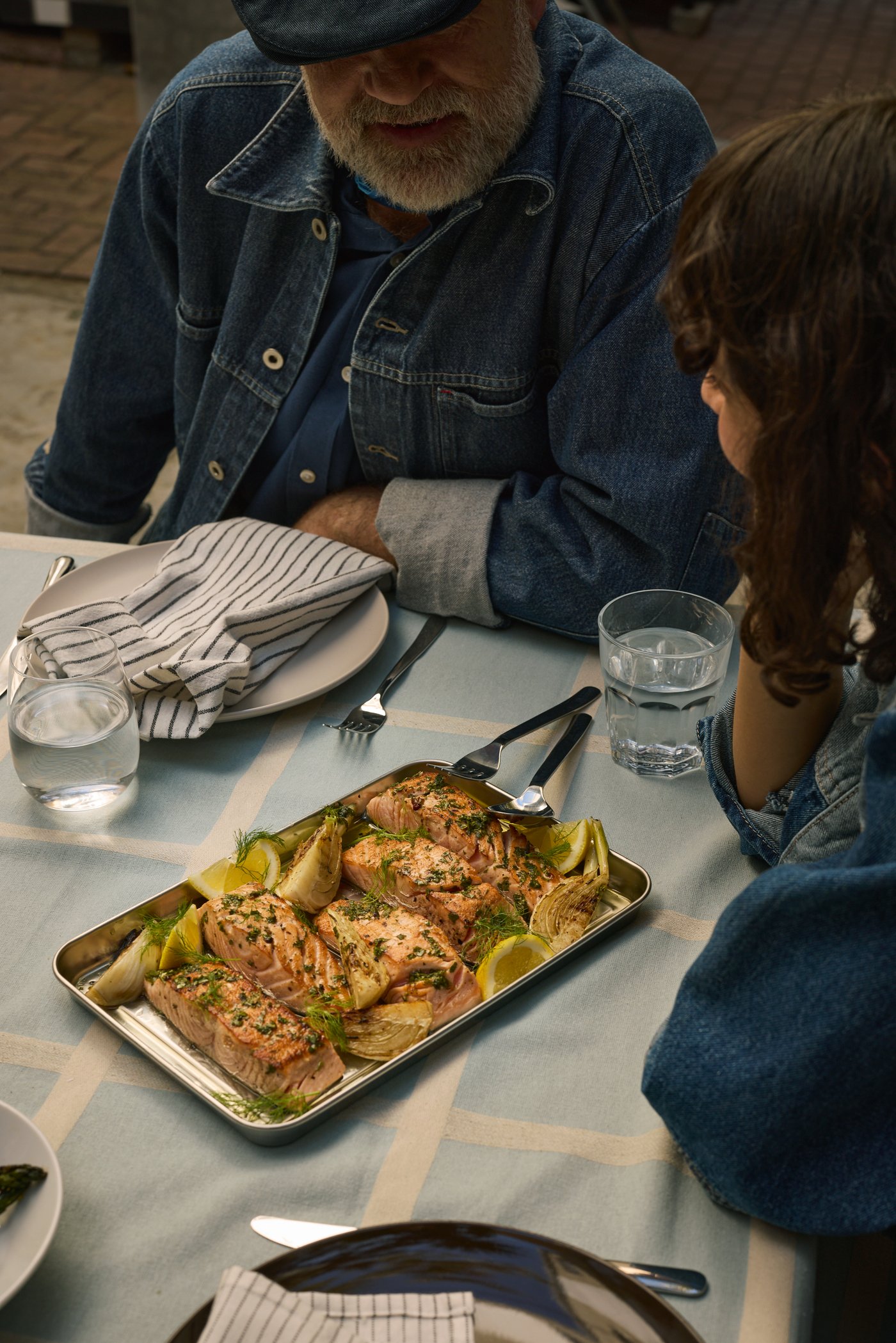 Auf einem Tisch liegt ein GRILLTIDER Blech mit einem leckeren Mittagsessen. Davor sitzten 2 Personen die am Tisch sitzten und auf das Blech schauen. Vor ihnen steht weißes Geschirr und ein Glas mit Wasser.