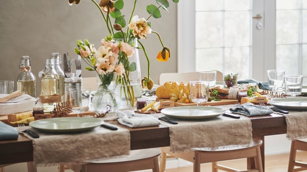 Una taula llarga MÖRBYLÅNGA parada per a una festa amb un teixit AINA, plats FÄRGKLAR, flors en gerros i diverses decoracions.