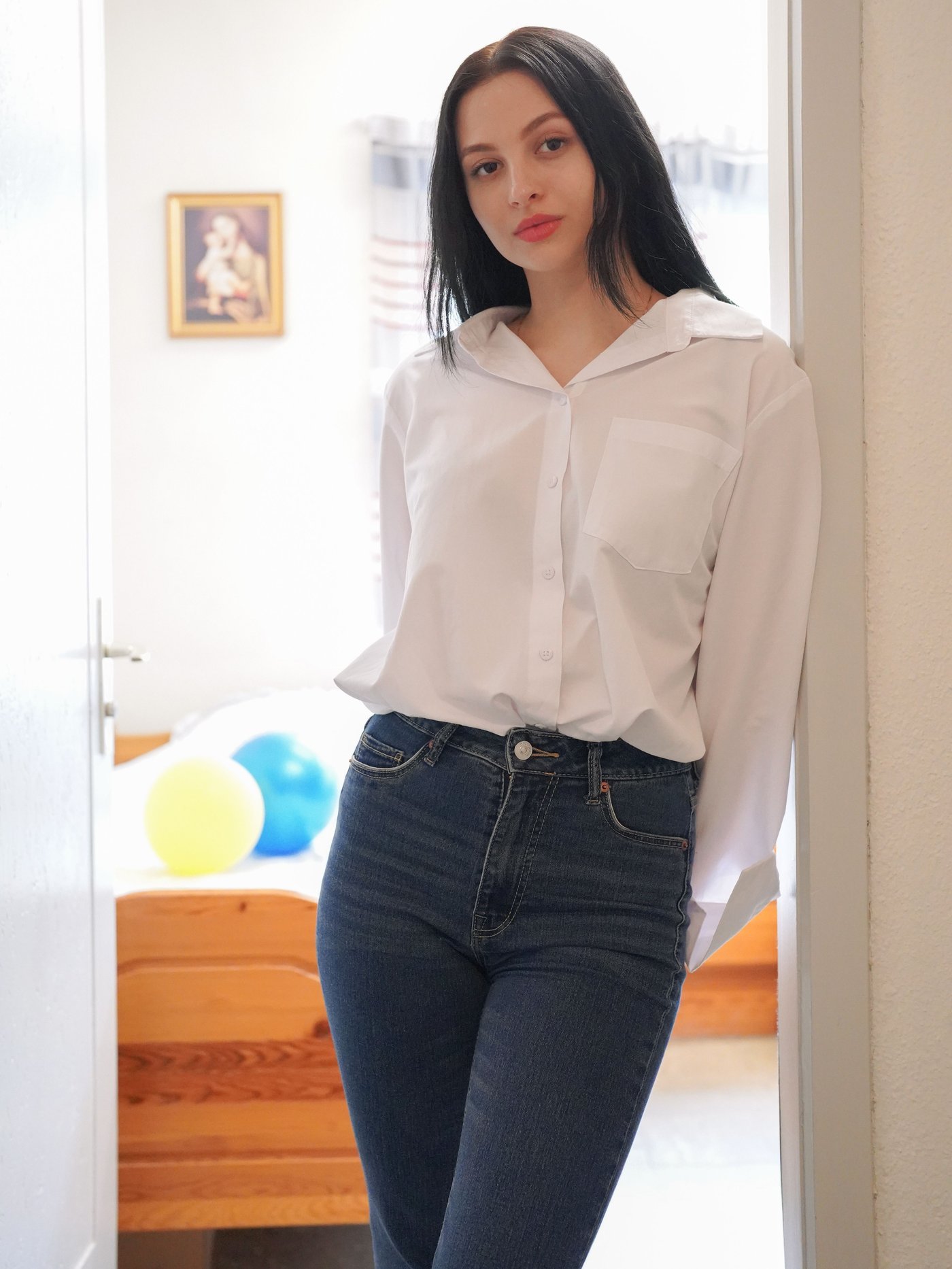 Girl leans against the doorframe in her living room and wears jeans and a white shirt.