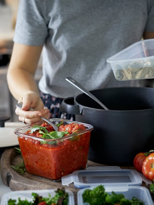 A person spoons tomato sauce from an IKEA 365+ square glass food container into a slim plastic container holding pasta.