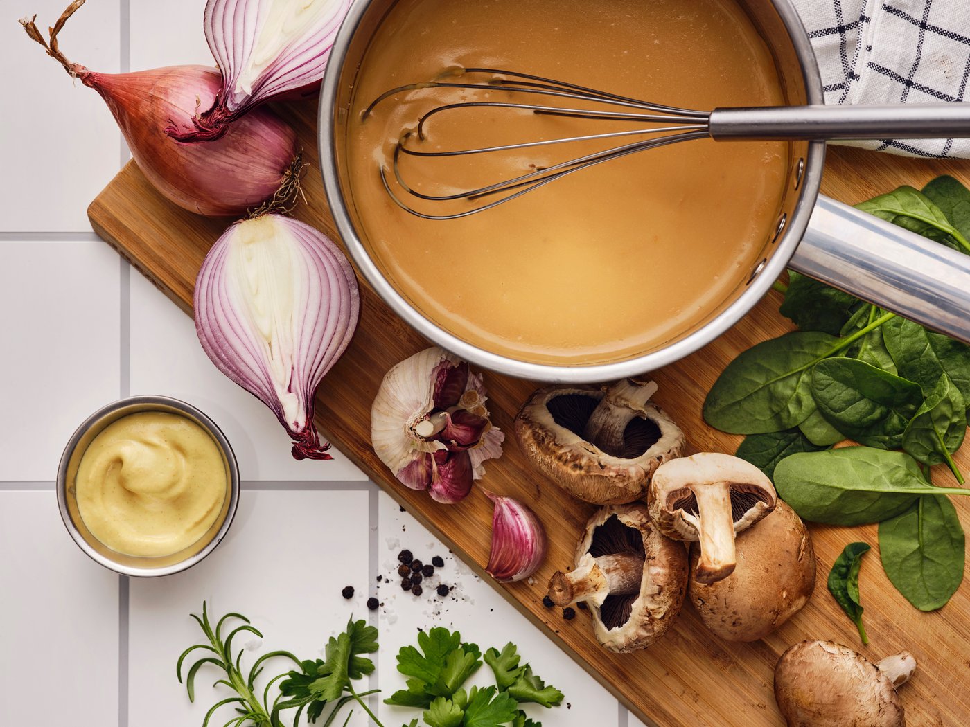 Sur une planche à découper où se trouvent de l’ail, des champignons, des herbes aromatiques et des échalotes, fouet dans une casserole contenant une préparation pour sauce ALLEMANSRÄTTEN.