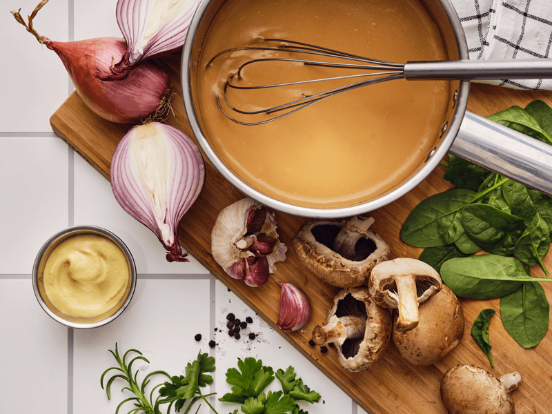 På et skjærebrett med hvitløk, sopp, urter og sjalottløk ligger en visp i en kasserolle med ALLEMANSRÄTTEN velsmakende sausblanding.