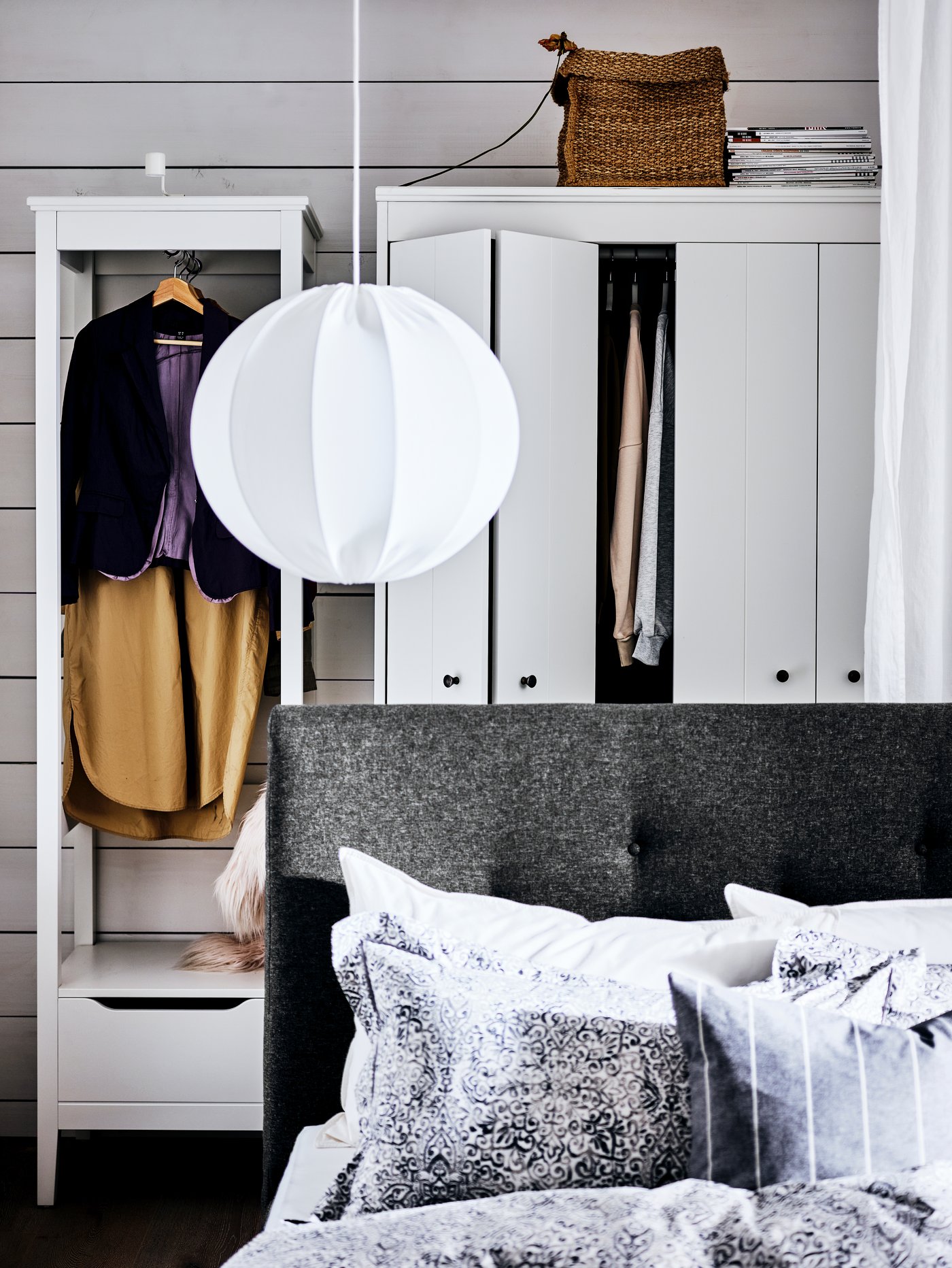 Cama tapizada IDANÄS gris con una lámpara de techo encima, frente a dos armarios blancos con ropa colgada.