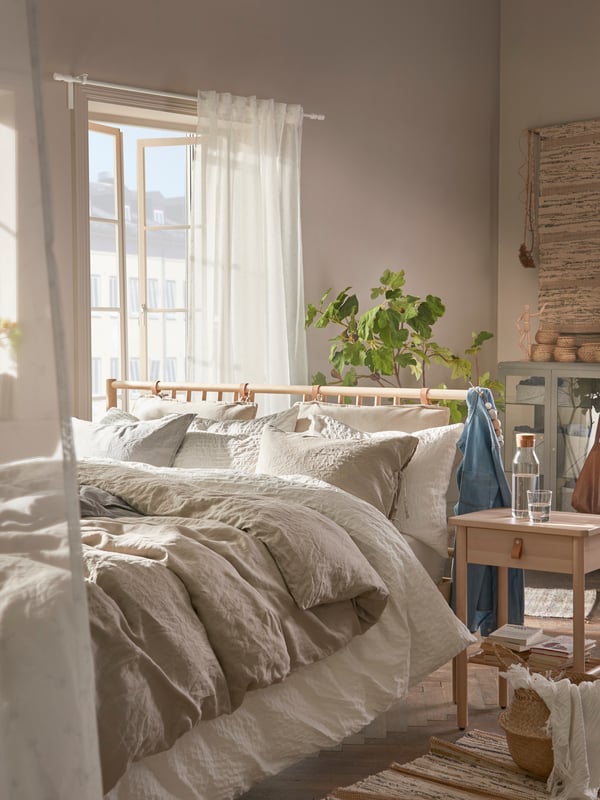 In einem Zimmer steht ein Bett in der Ecke und dahinter ein kleiner Kasten mit einer Vase darauf. Das bett hat eine dunkle Decke und weiße Pölster.