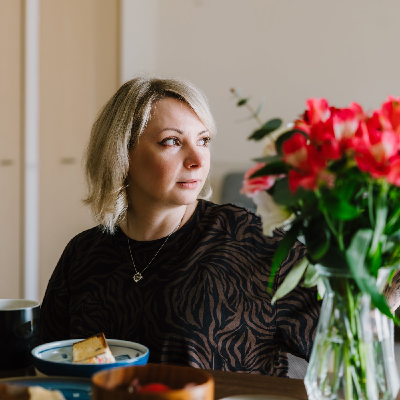 A blonde woman sitting by a kitchen table looking to the right, a vase with red roses stands next to her.