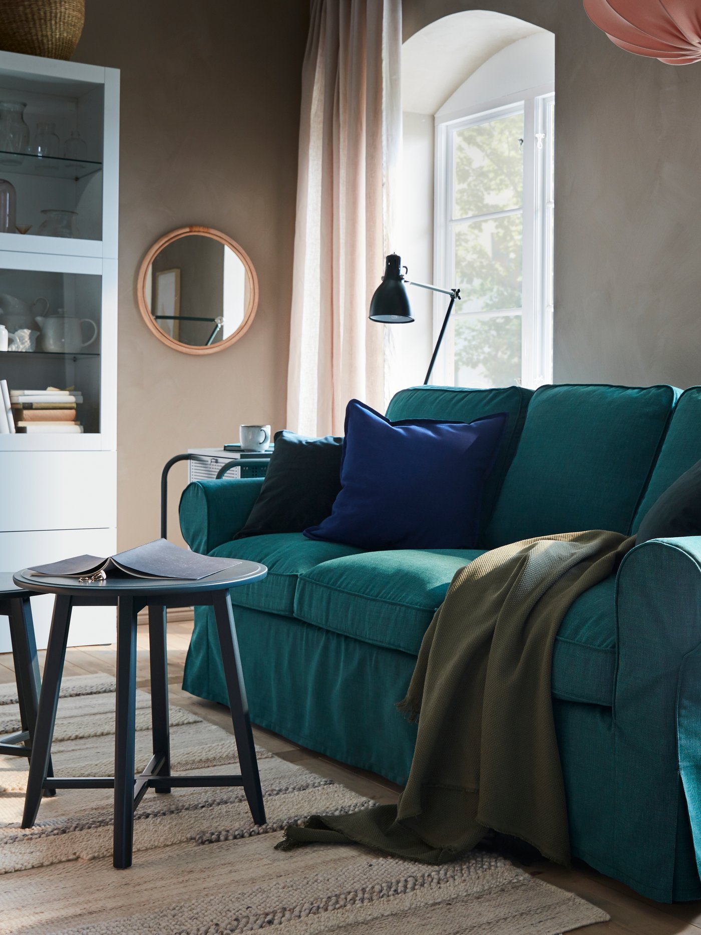 A sofa in dark turquoise, a small table in dark blue-green, a green throw, a striped rug and a pink pendant lamp.