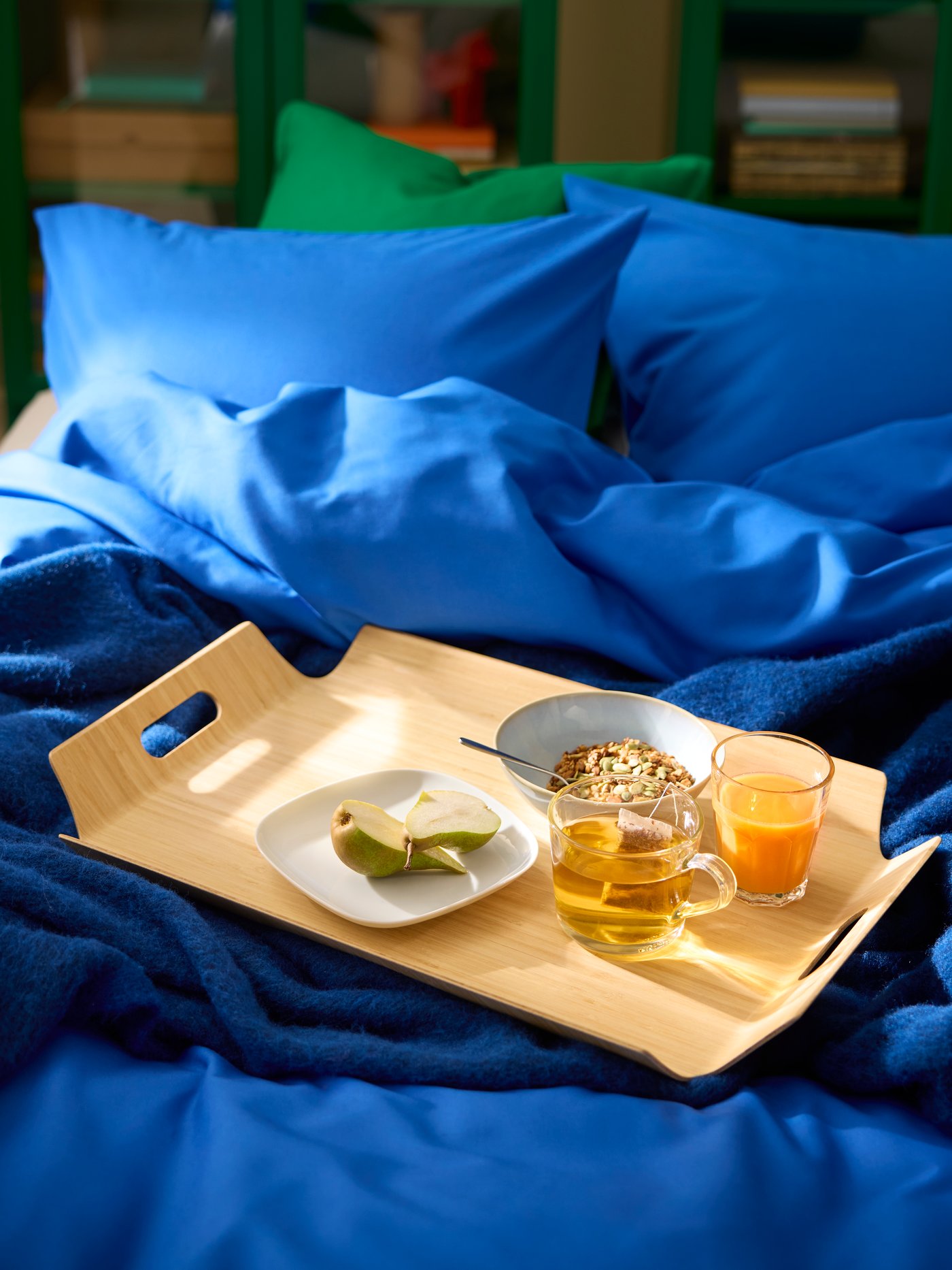 Morgenmad på lys træbakke med håndtag på seng med blåt sengetøj.