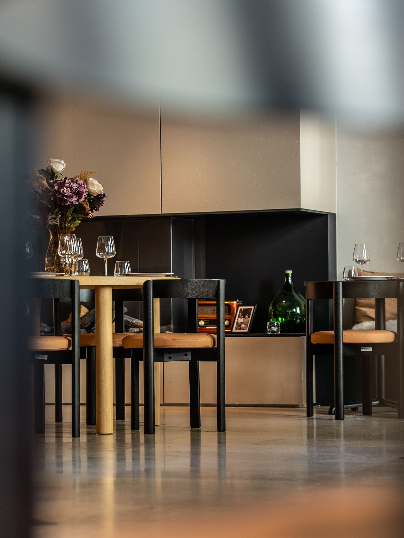 Moderne eetruimte met houten tafel, zwarte stoelen, bloemenvaas, wijnglazen en decoratieve kast op gepolierde betonvloer. Warme, gezellige sfeer.