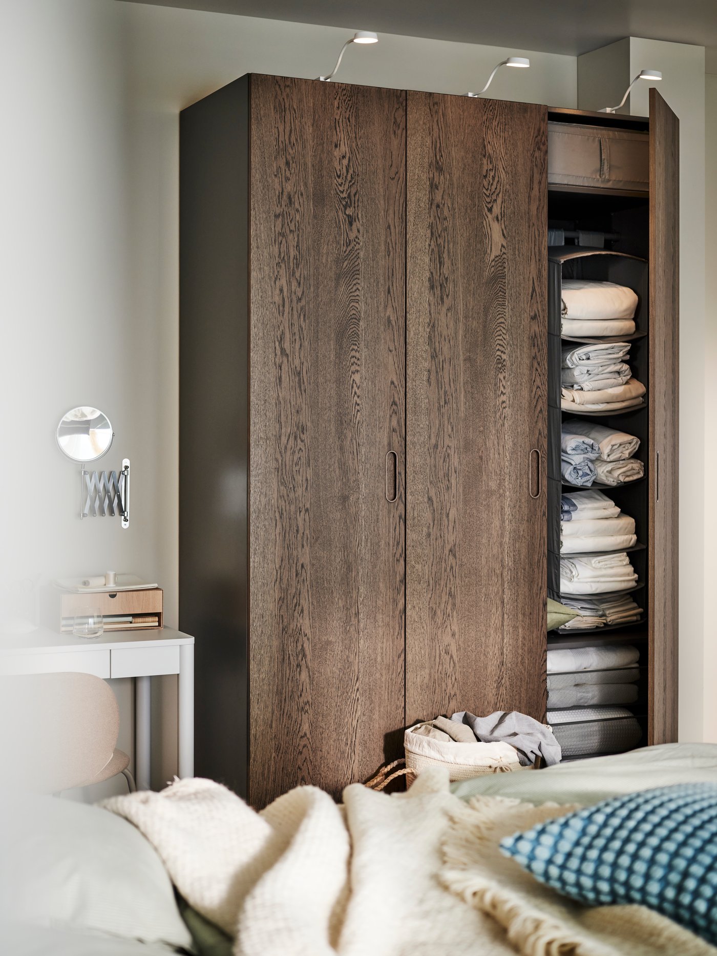 PAX/TONSTAD wardrobe in brown stained oak veneer, with one door open showing neatly folded sheets. Bed in foreground.