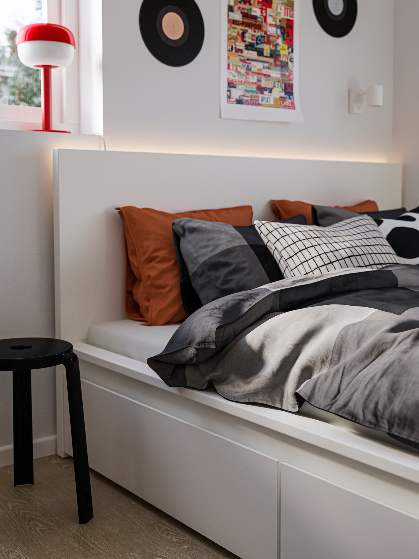 Nahaufnahme eines weißen MALM-Bettes mit Schubladen, BRUNKRISSLA-Bettwäsche, einem schwarzen Hocker an der Seite und einer roten Lampe im Fenster.