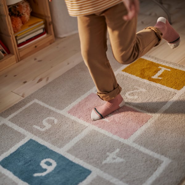A child is hopping on a beige STÄPPSPARV rug in a sunny bedroom with a light white stained pine TROFAST storage unit nearby.