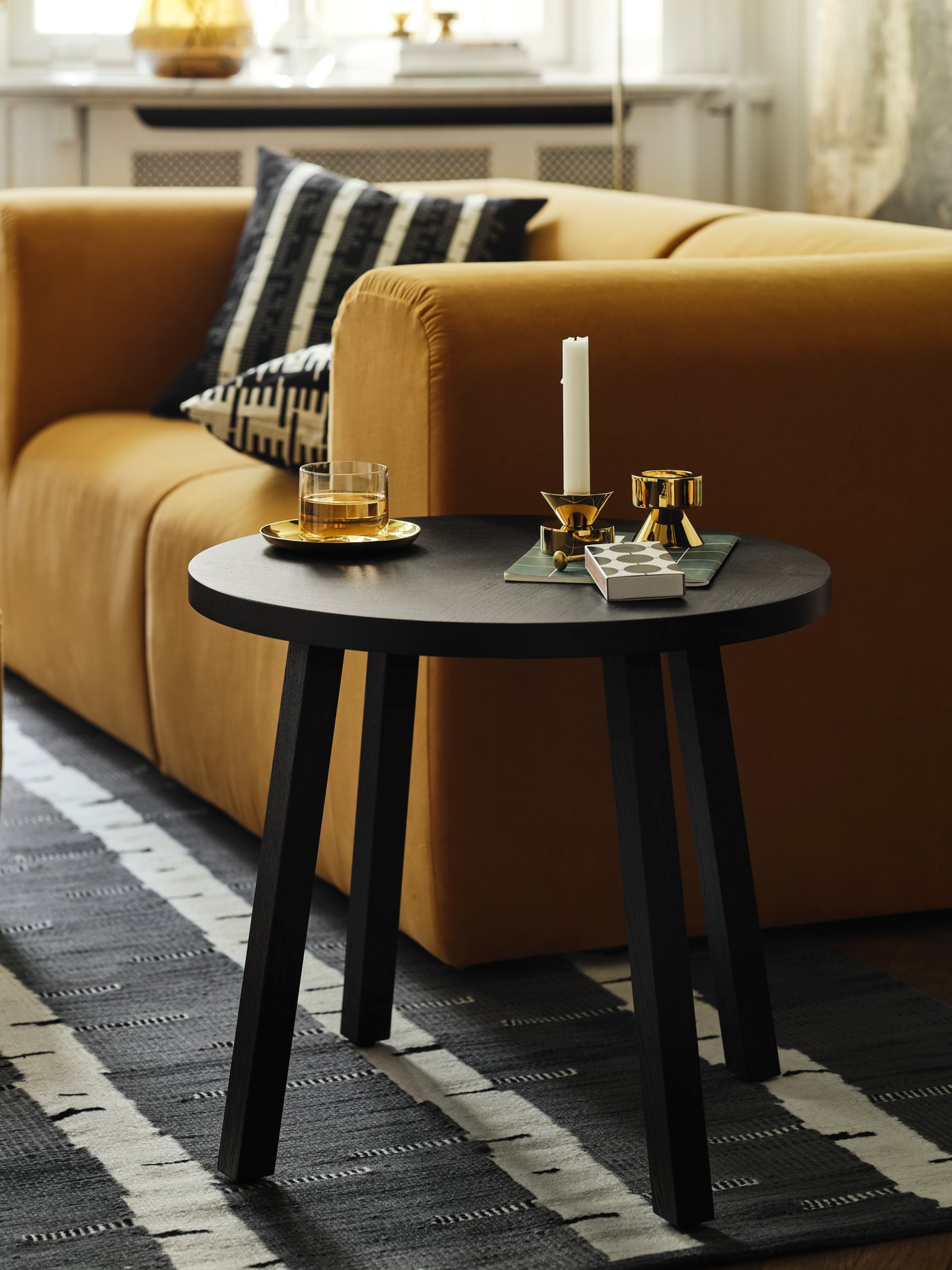 Runt svart sidobord bredvid en gul soffa, dekorerat med ljusstake och små föremål.