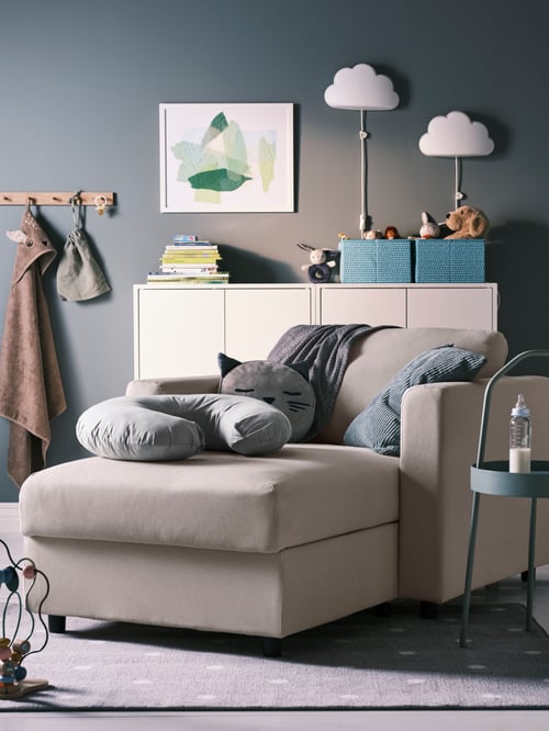 Bedroom with a grey nursing pillow on a footstool, wall-mounted shelving units, a baby towel on a rack, and a side table.