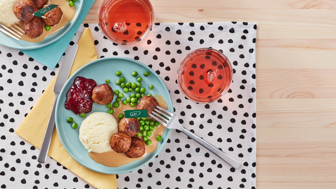 Assiette divisée montrant des boulettes de viande avec des pois verts et de la purée de pommes de terre, accompagnées de sauce et de confiture sur un plateau blanc, à côté d'une table avec une salade, des tomates cerises et un emballage d'IKEA.