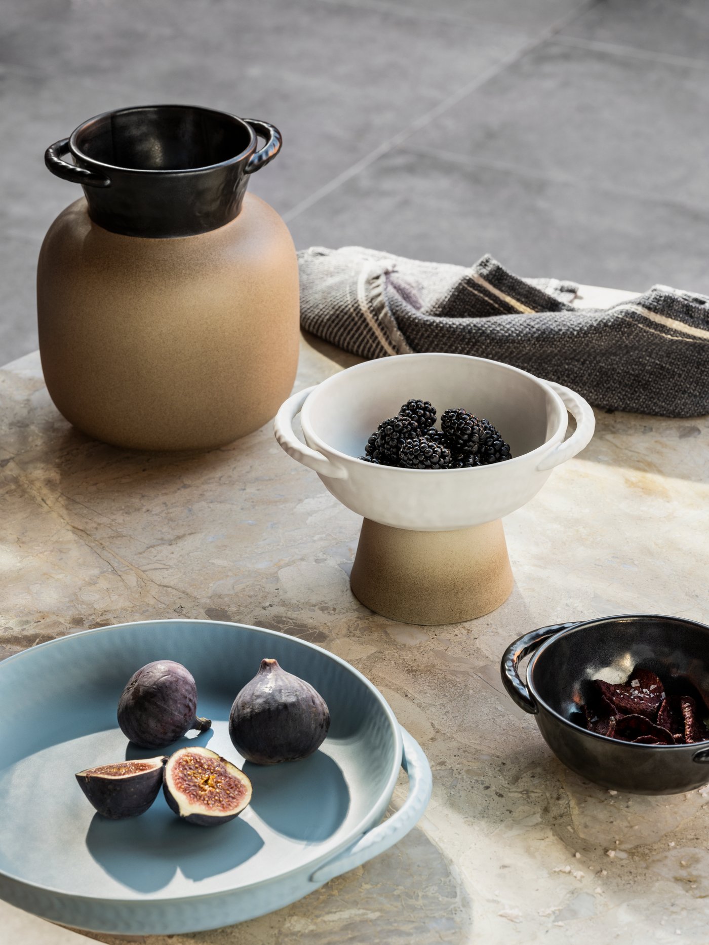 A vase, a plate and two bowls from the LOKALT collection placed on a marble table top against a gray background.