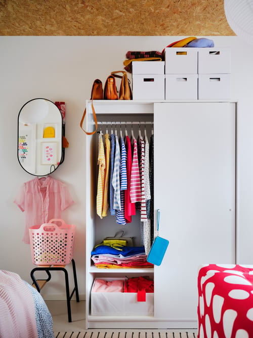 Les portes coulissantes à moitié ouvertes d’une armoire-penderie blanche révèlent des vêtements colorés et des boîtes blanches sur le dessus.