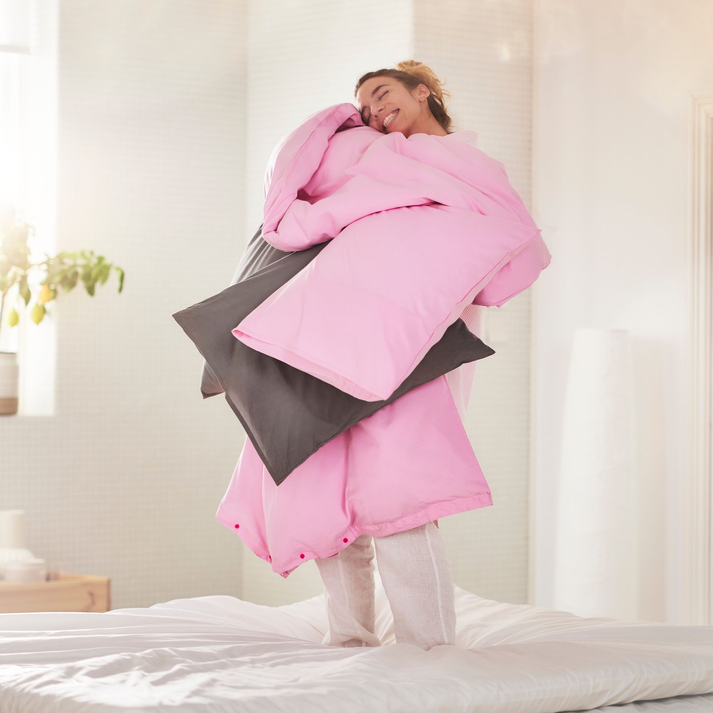 A person is smiling as they hold a duvet and pillows in light pink PILTANDVINGE bed linen to them a light bedroom.