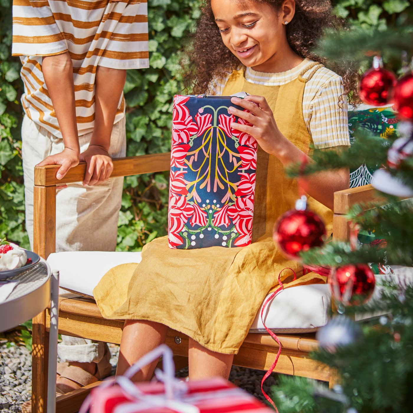 Una niña abriendo un regalo de Navidad en un jardín y detrás de ella un árbol de navidad.