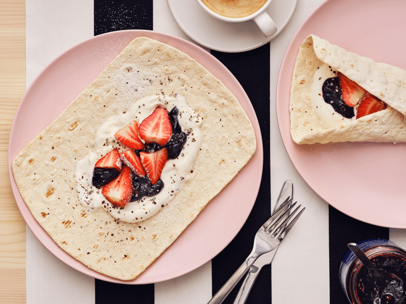 In un piatto rosa, appoggiato su una superficie a righe bianche e nere, una fetta di pane sottile con frutti di bosco e panna montata.