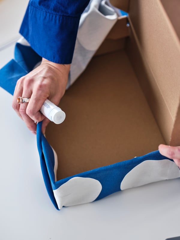 A person glues blue/white RAKLOSTA pre-cut fabric onto a VATTENTRÅG box with lid to give the box a colourful cover.