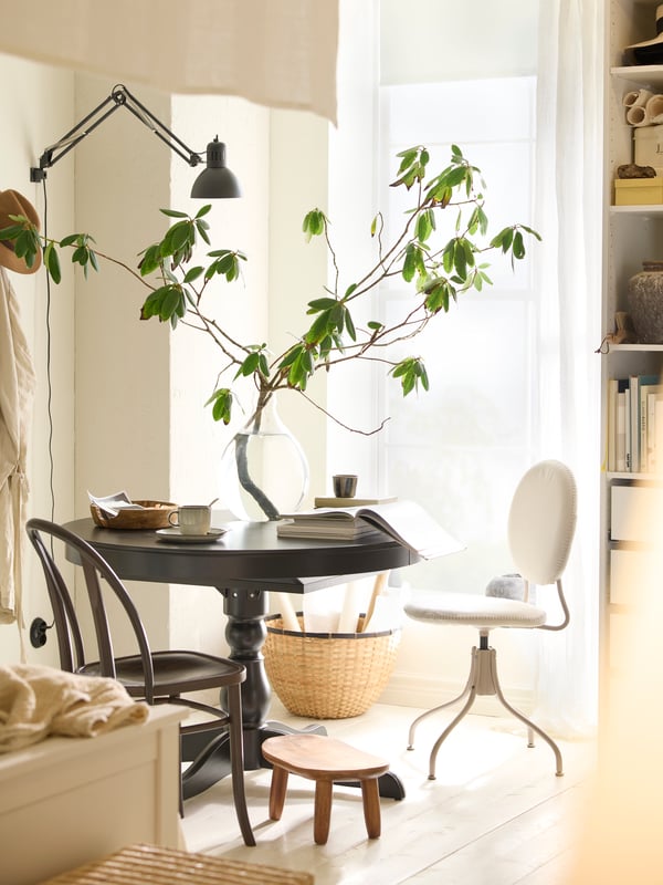 A black INGATORP dining table by a window with a glass vase on it and a beige BJÖRKBERGET swivel chair and stool in front.