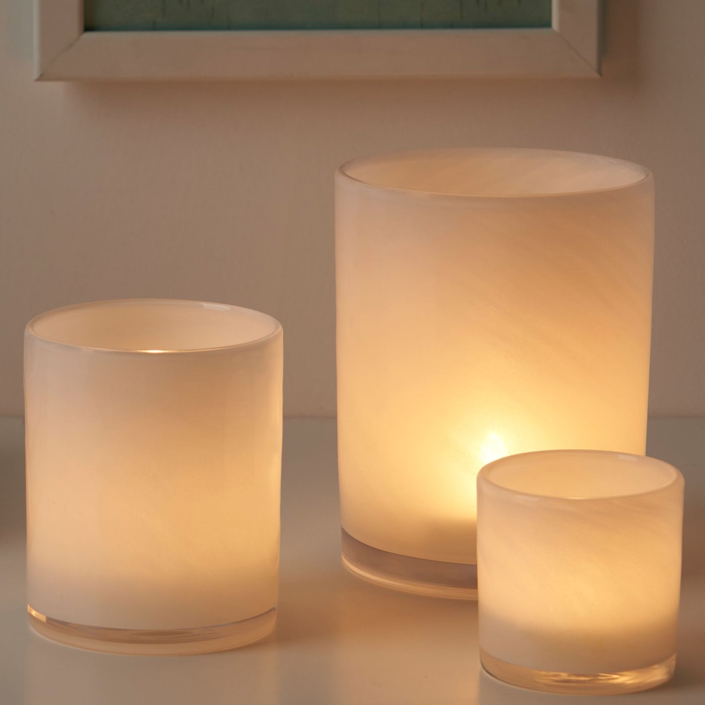 Tre stycken vita VINDSTILLA ljuslyktor i tre olika storlekar med tända ljus som sprider ett varmt sken. 