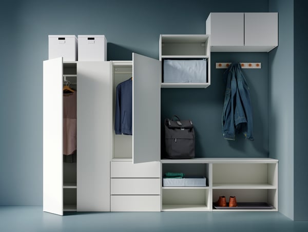 Planificador PLATSA de color blanco con diferentes divisiones y tamaños, colgadores en su pared con chaqueta colgada, mochila sobre el mueble y cajas en la parte superior.